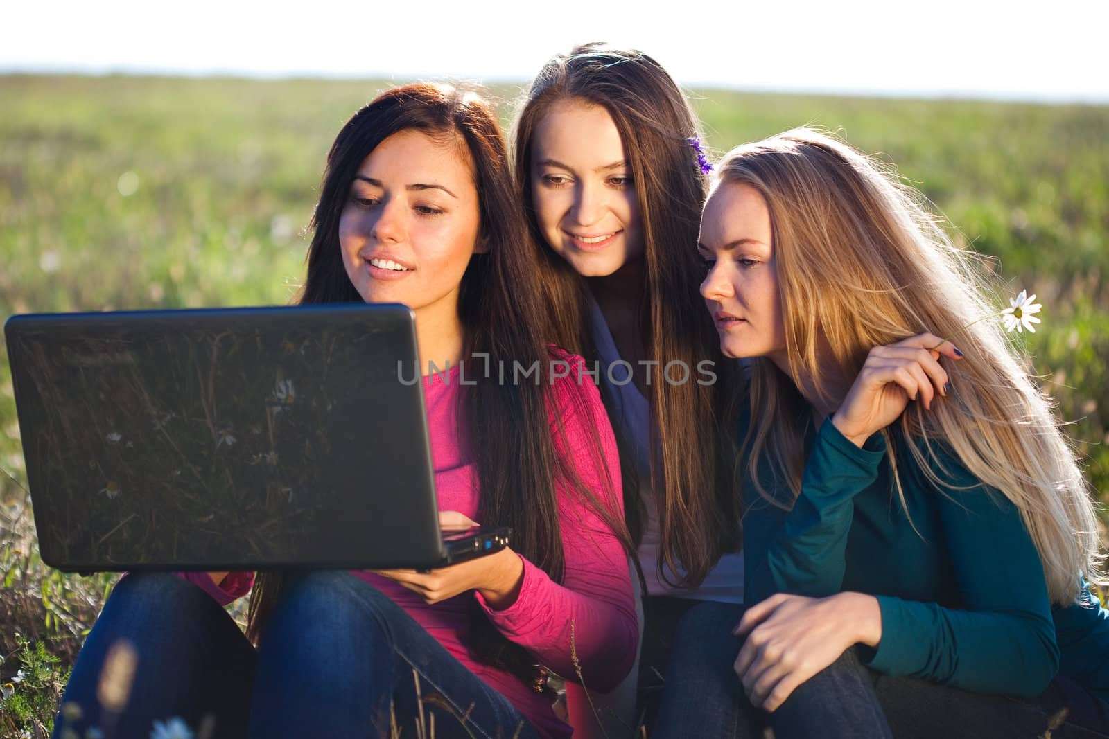 three young beautiful woman with a laptop sitting in the field on sky background 