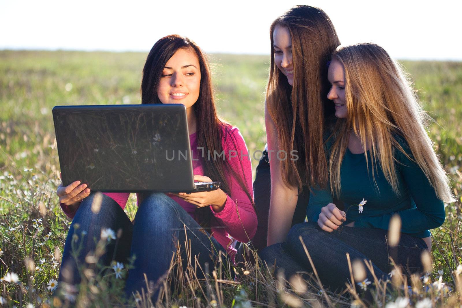 three young beautiful woman with a laptop sitting in the field o by jannyjus