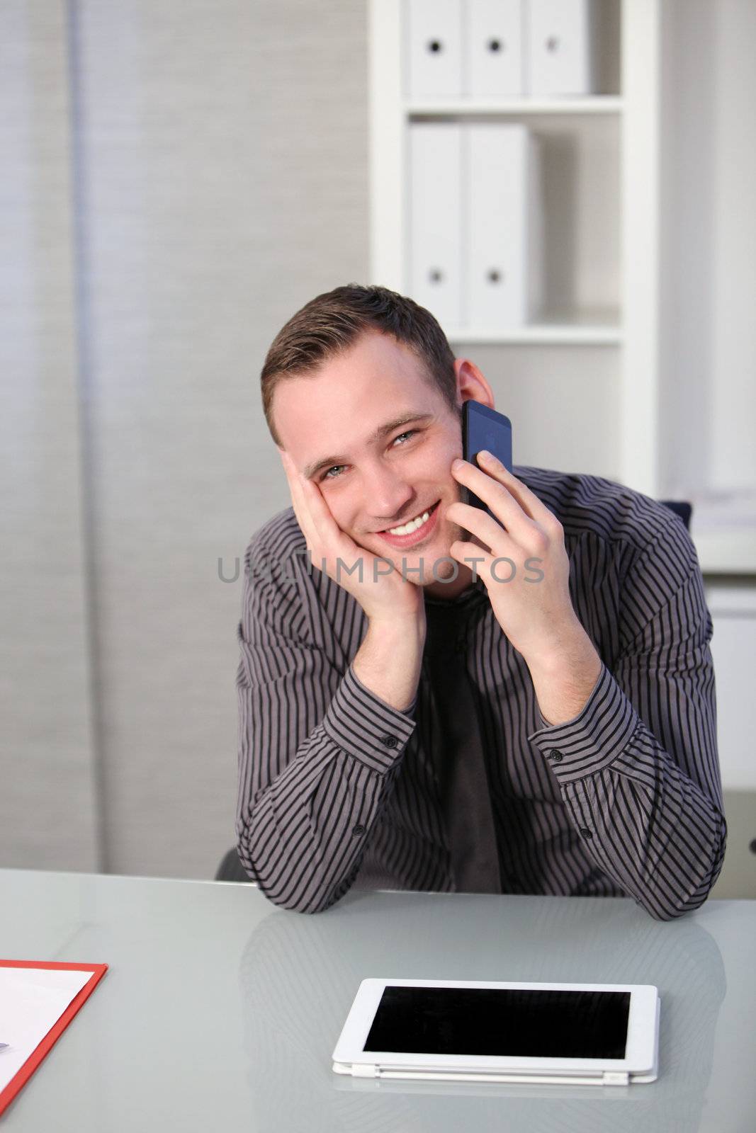Handsome young business man using a mobile while seated at his desk as he looks up to smile at the camera