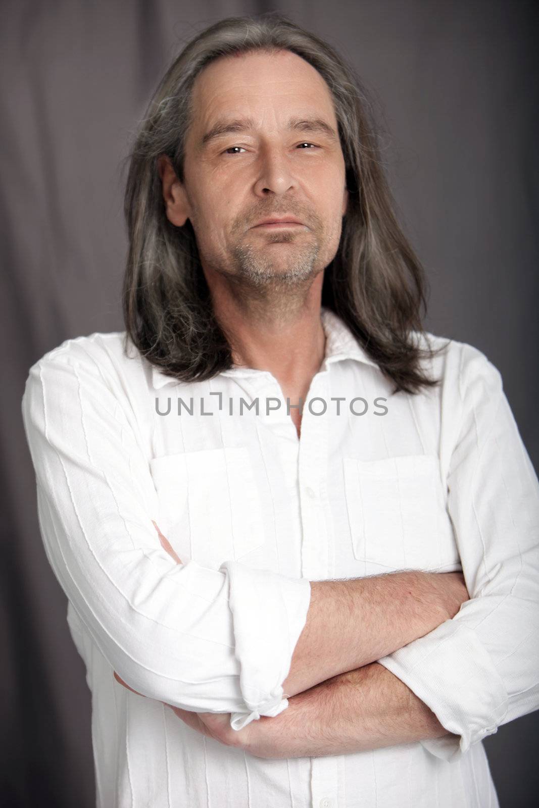 Sexy macho middle-aged man with shoulder length hair and a stubbly beard standing with his arms folded and an amused expression looking at the camera