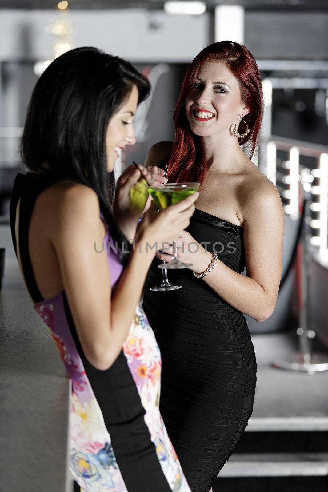 Two beautiful young woman chatting and drinking cocktails at a nightclub or wine bar.