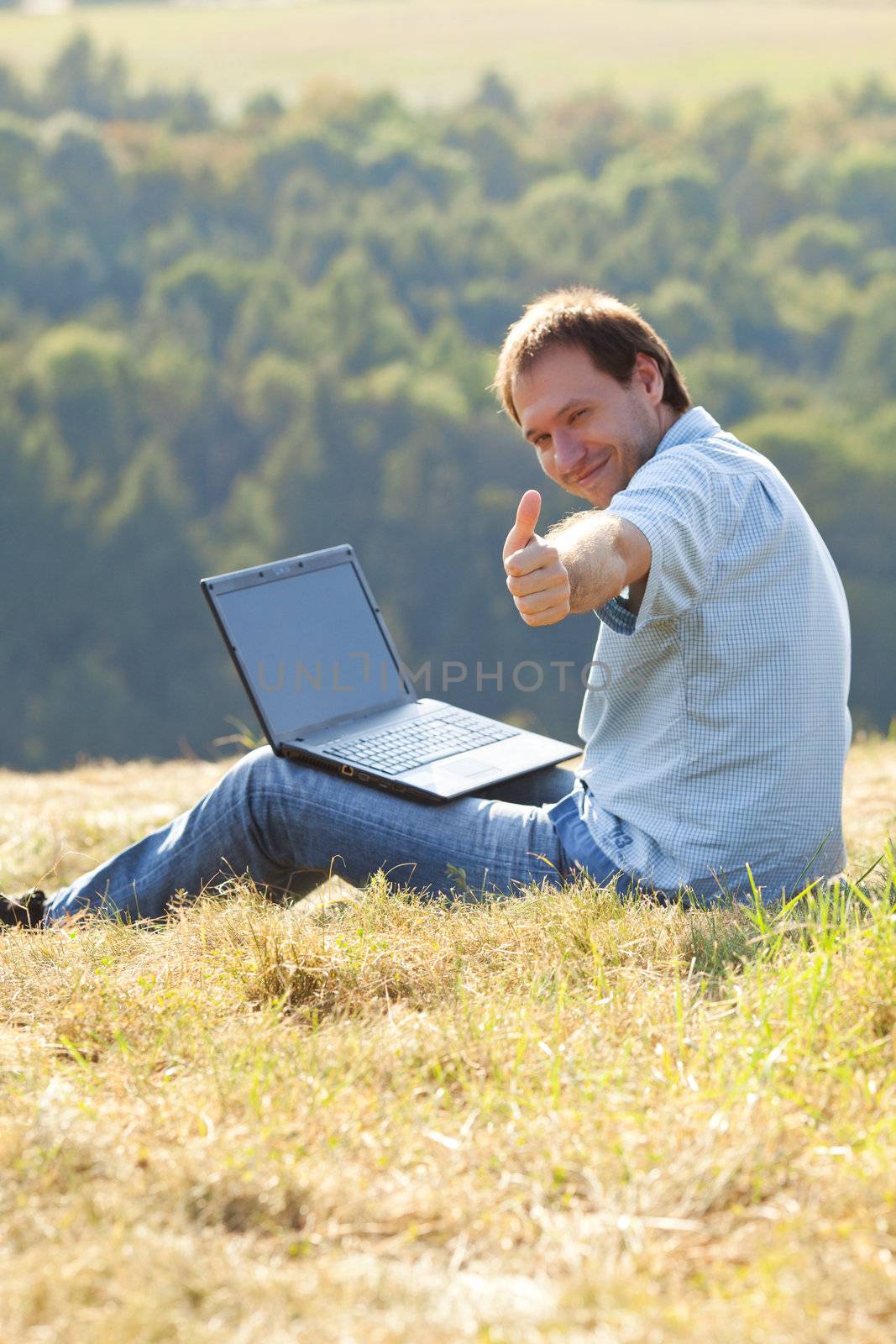 young man using laptop sitting on the grass on the hillside by jannyjus