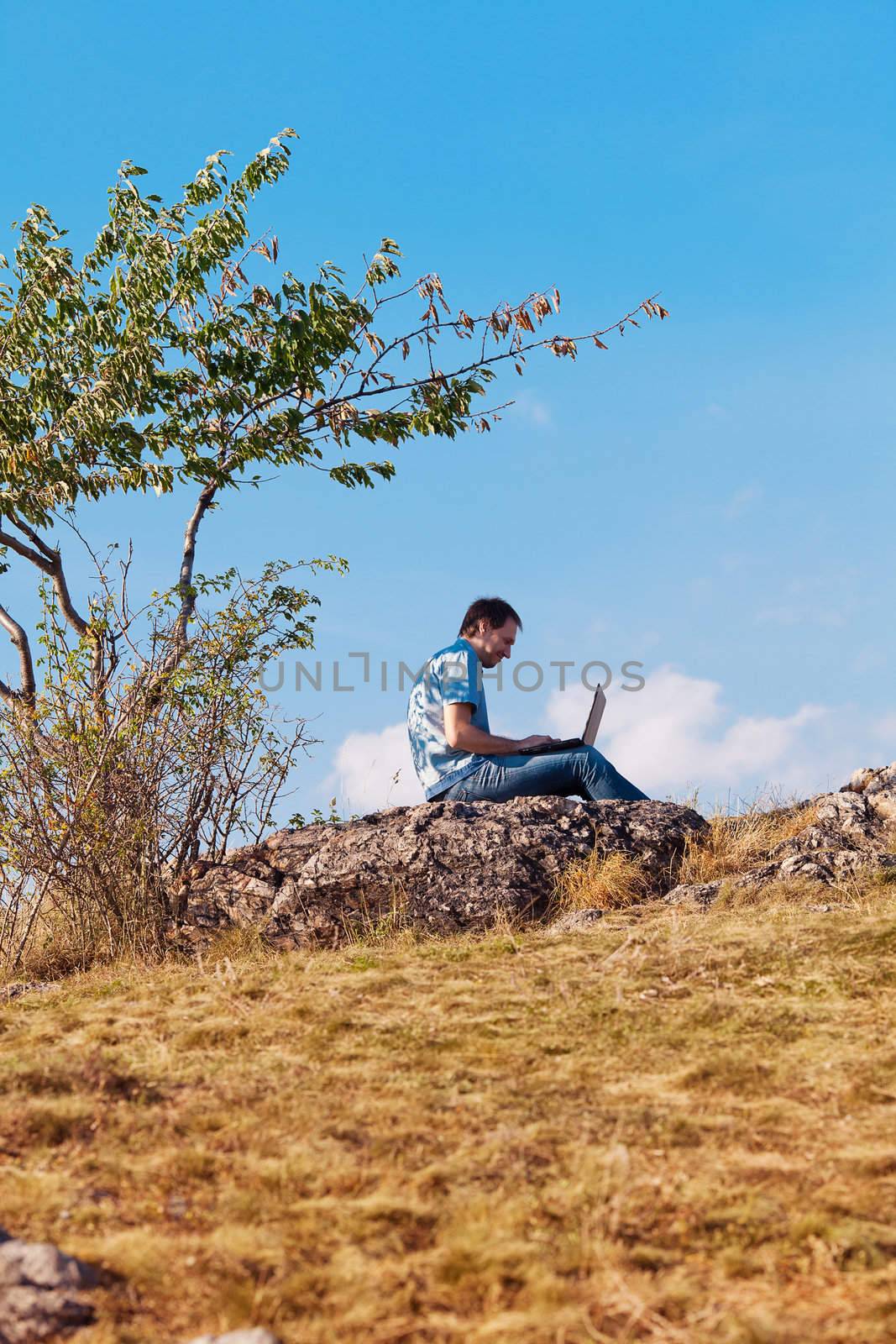 young man using laptop sitting on a hill against the blue sky