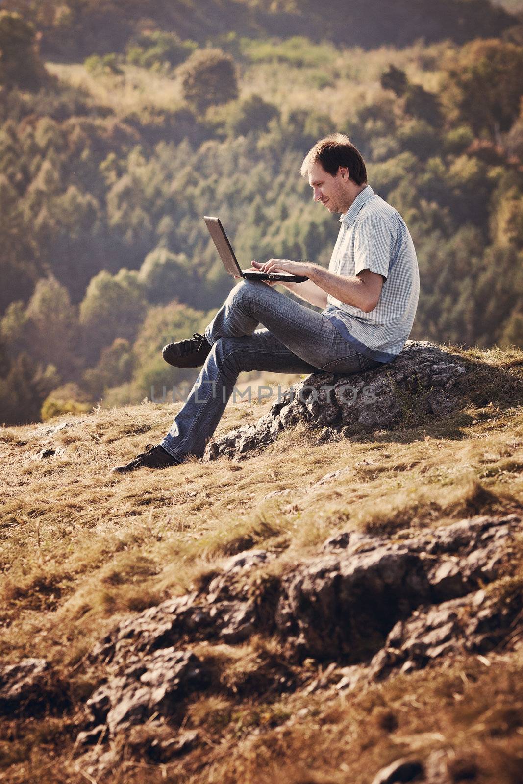young man using laptop sitting on the grass on the hillside by jannyjus