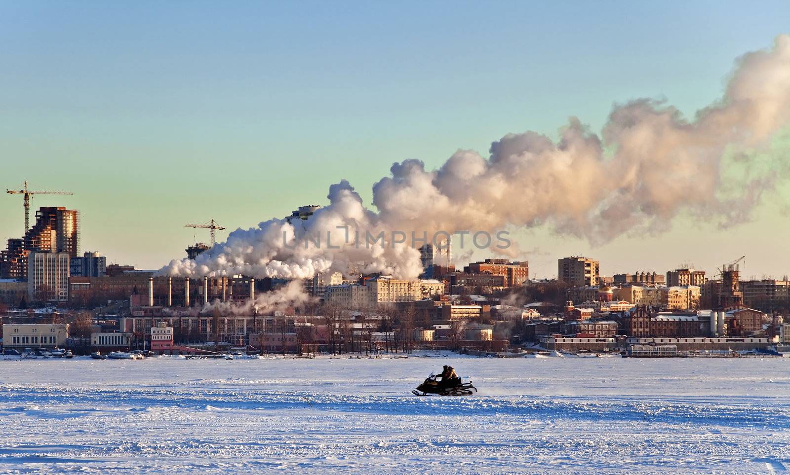 Snowmobile rides on the frozen river in the background of the city with the industrial landscape. Sunset. Samara. Russia.