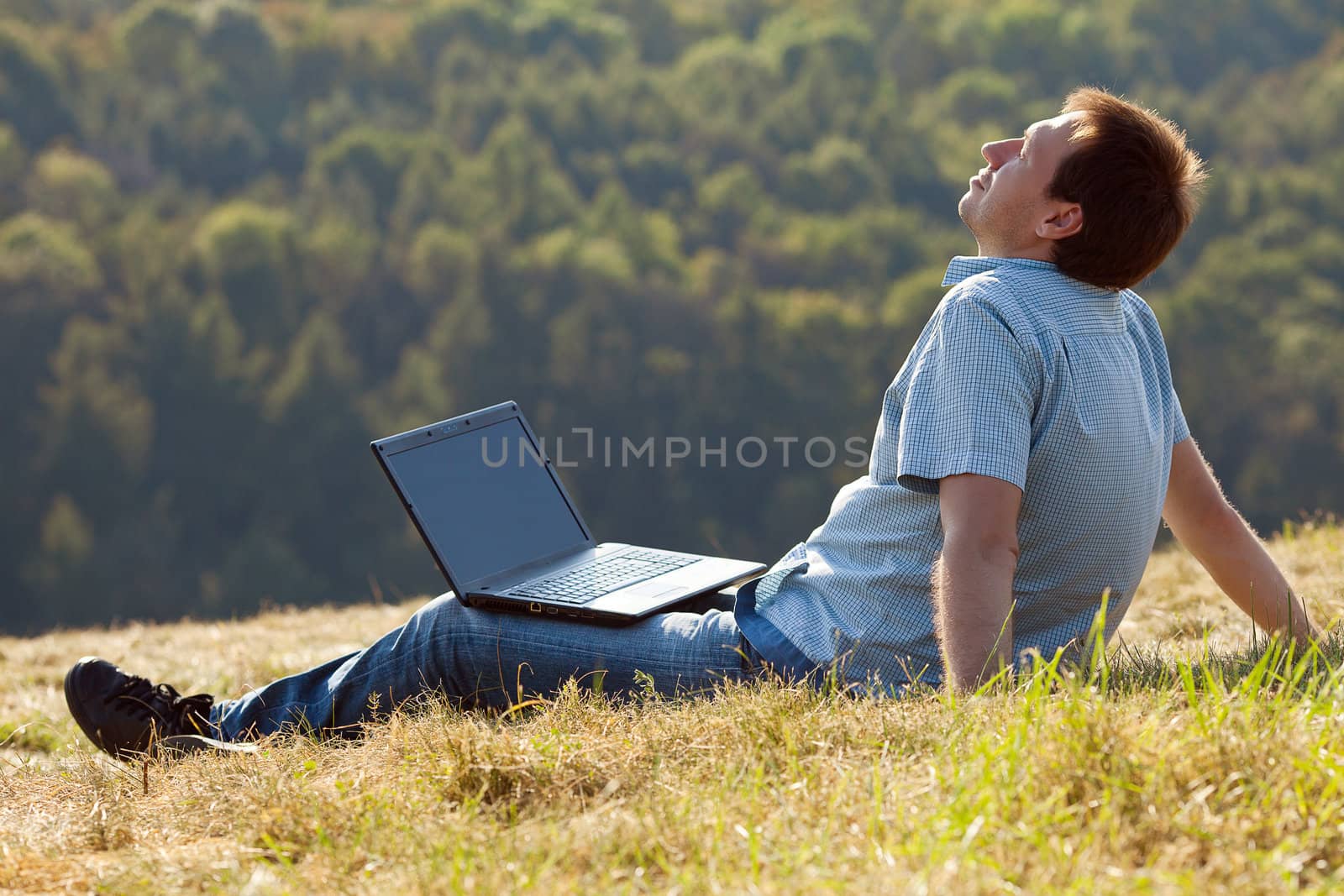 young man using laptop sitting on the grass on the hillside by jannyjus