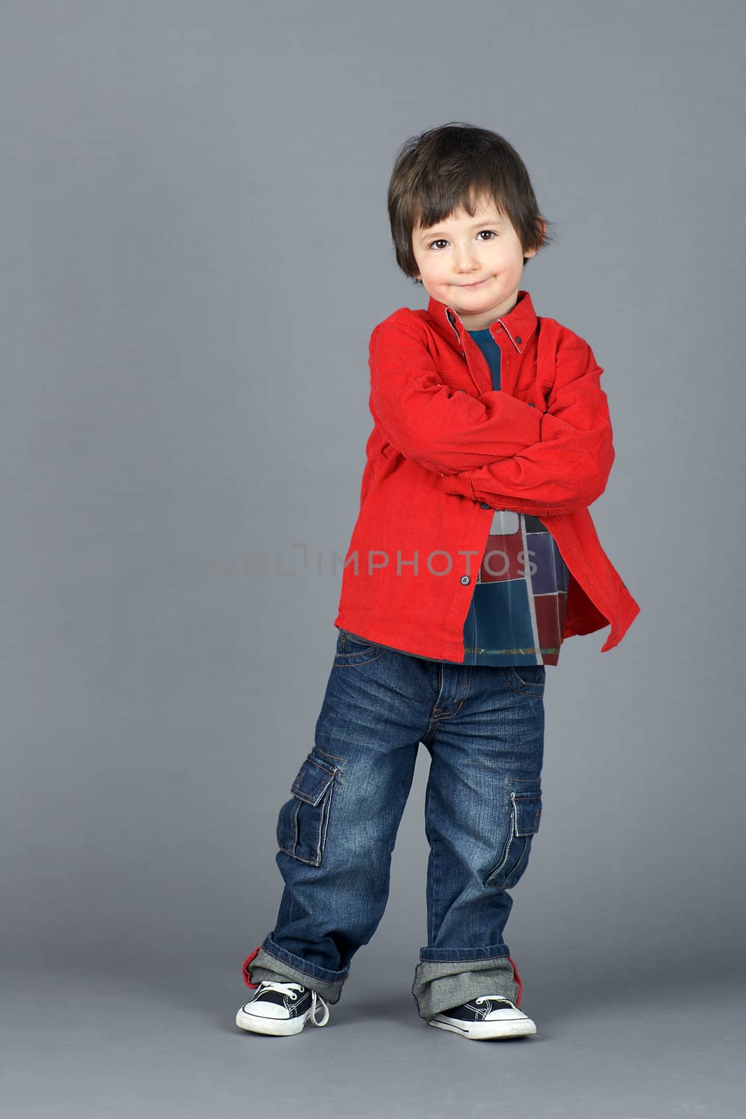 Super cute young or little boy smiling dressed in cool red corduroy shirt and jeans, leaning on one side, shot in studio  over grey background.