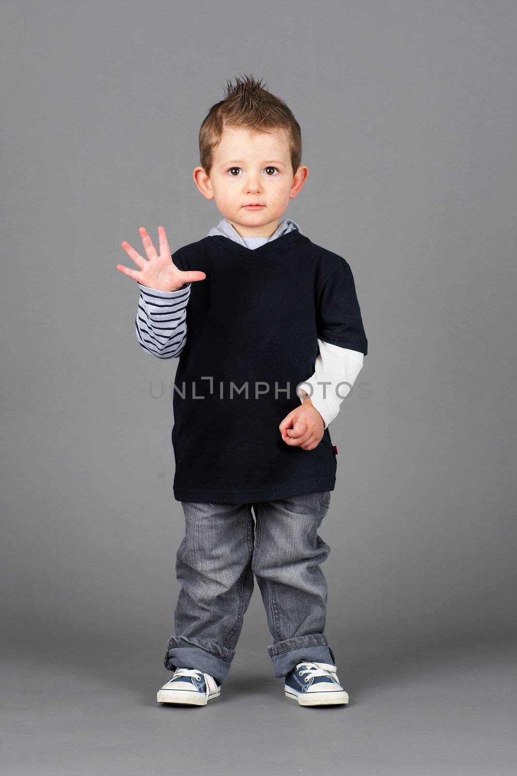 Cute and hip little boy wearing jeans and running shoes waving hello or showing fingers to count, shot in studio over grey background.