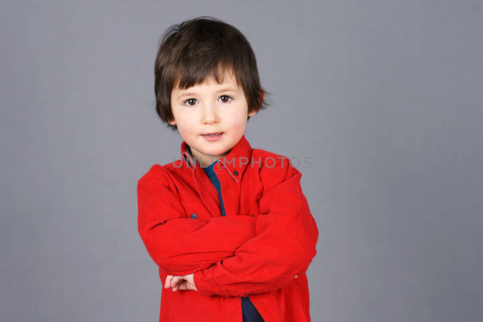 Cute little boy with little smile big brown eyes and crossed arms dressed in red shirt, studio shot over grey background.