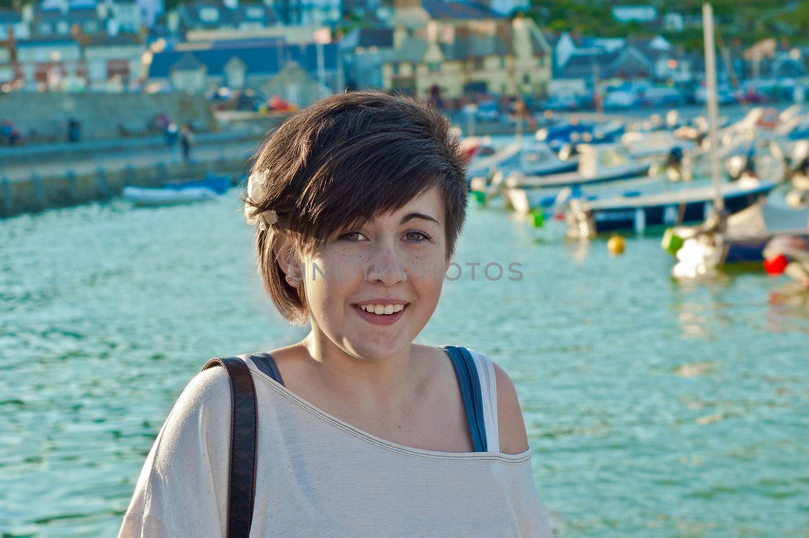 an image of a pretty young teenage girl looking towards you with a dock full of yatches, boats and buildings behind.
