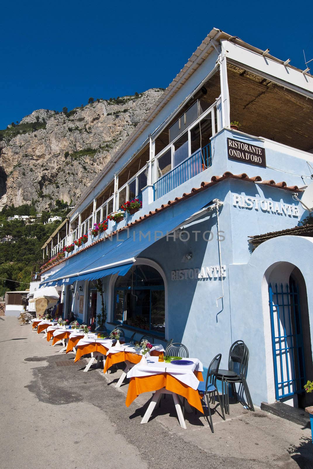 Brightly coloured tables outside blue cafe, under craggy mountains, Capri Italy