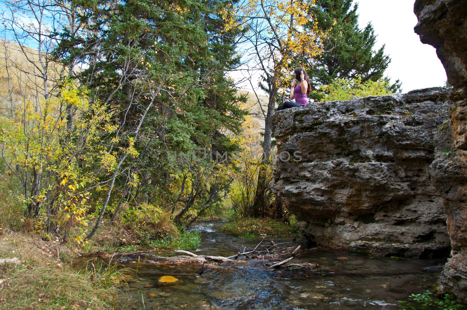 Young attractive girl sitting on the edge of a rock in autumn doing yoga and meditating with a creek running beneath her