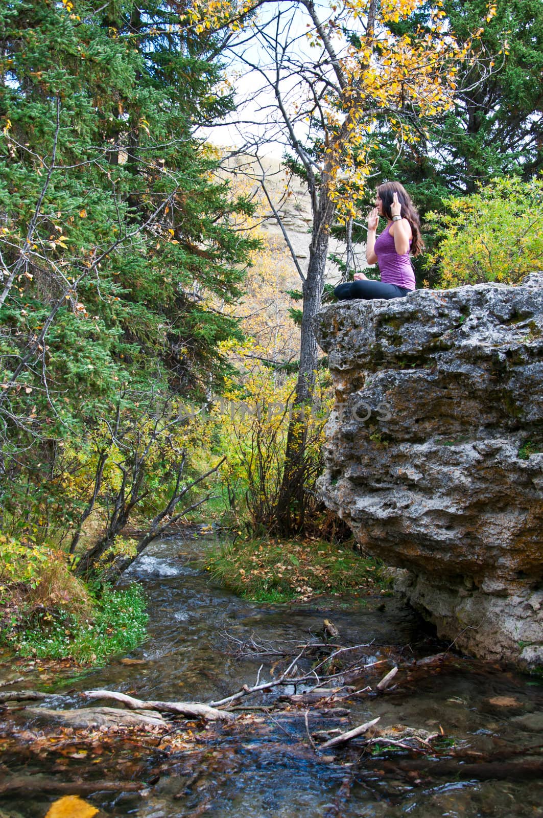 Young attractive girl sitting on the edge of a rock in autumn doing yoga and meditating with a creek running beneath her