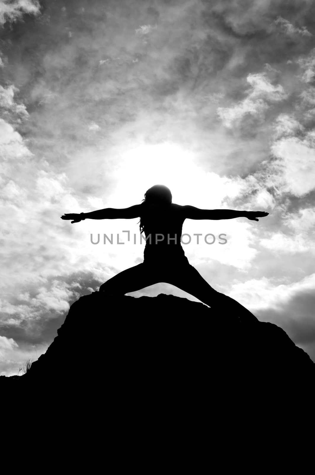 Black and white silhouette of a young attractive girl doing the Warriors pose from yoga on top of a rock with the sky and clouds behind her.
