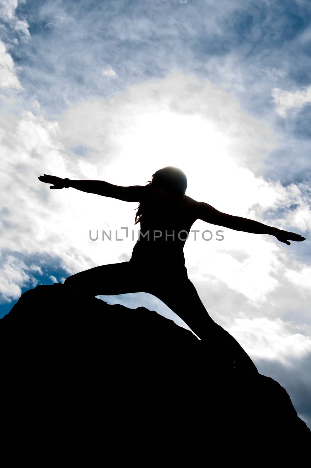 Angled silhouette of a young attractive girl doing the Warriors pose from yoga on top of a rock with the sky and clouds behind her.