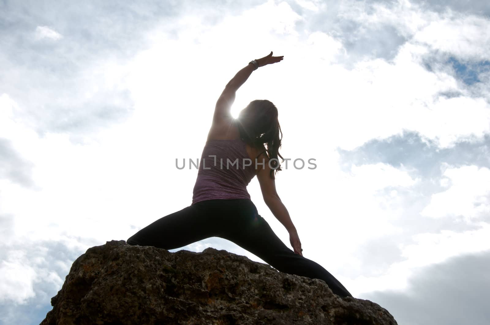 Silhouette of young attractive girl doing yoga on top of a rock with the sky and cloudy in the background
