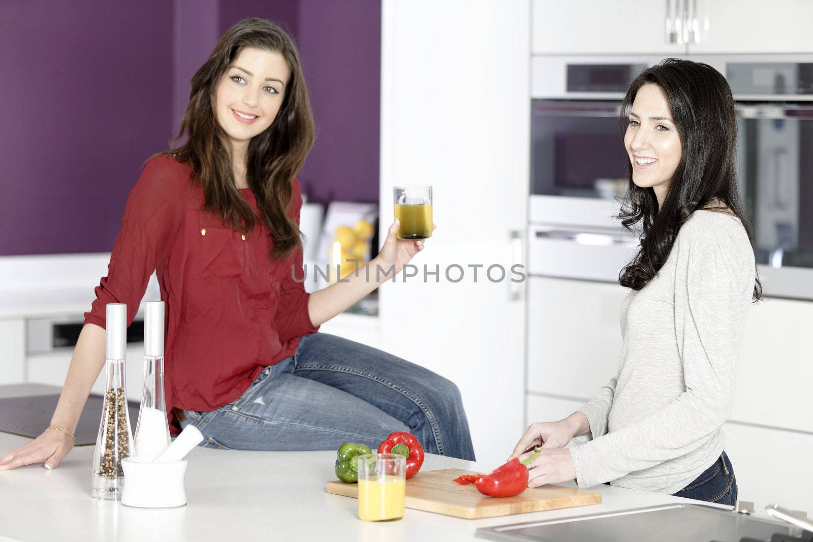 Two attractive young women preparing food in a white kitchen while talking.