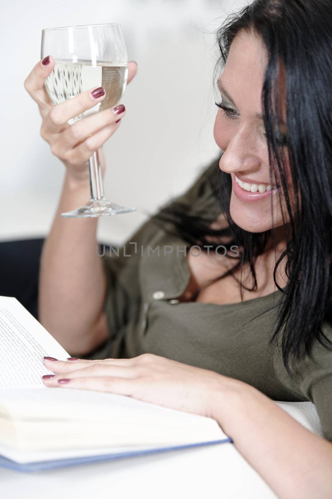 Attractive young woman lying on her sofa enjoying a glass of wine and a good book.