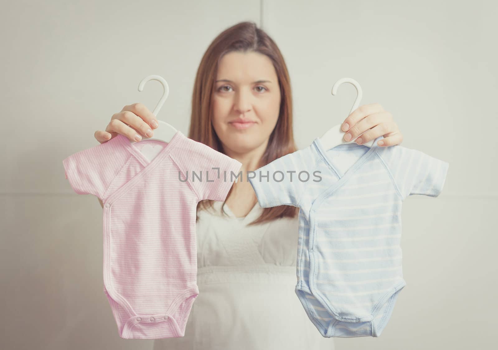 Beautiful pregnant young woman holding up bodysuits  for a girl or a boy newborn in soft pastel colors