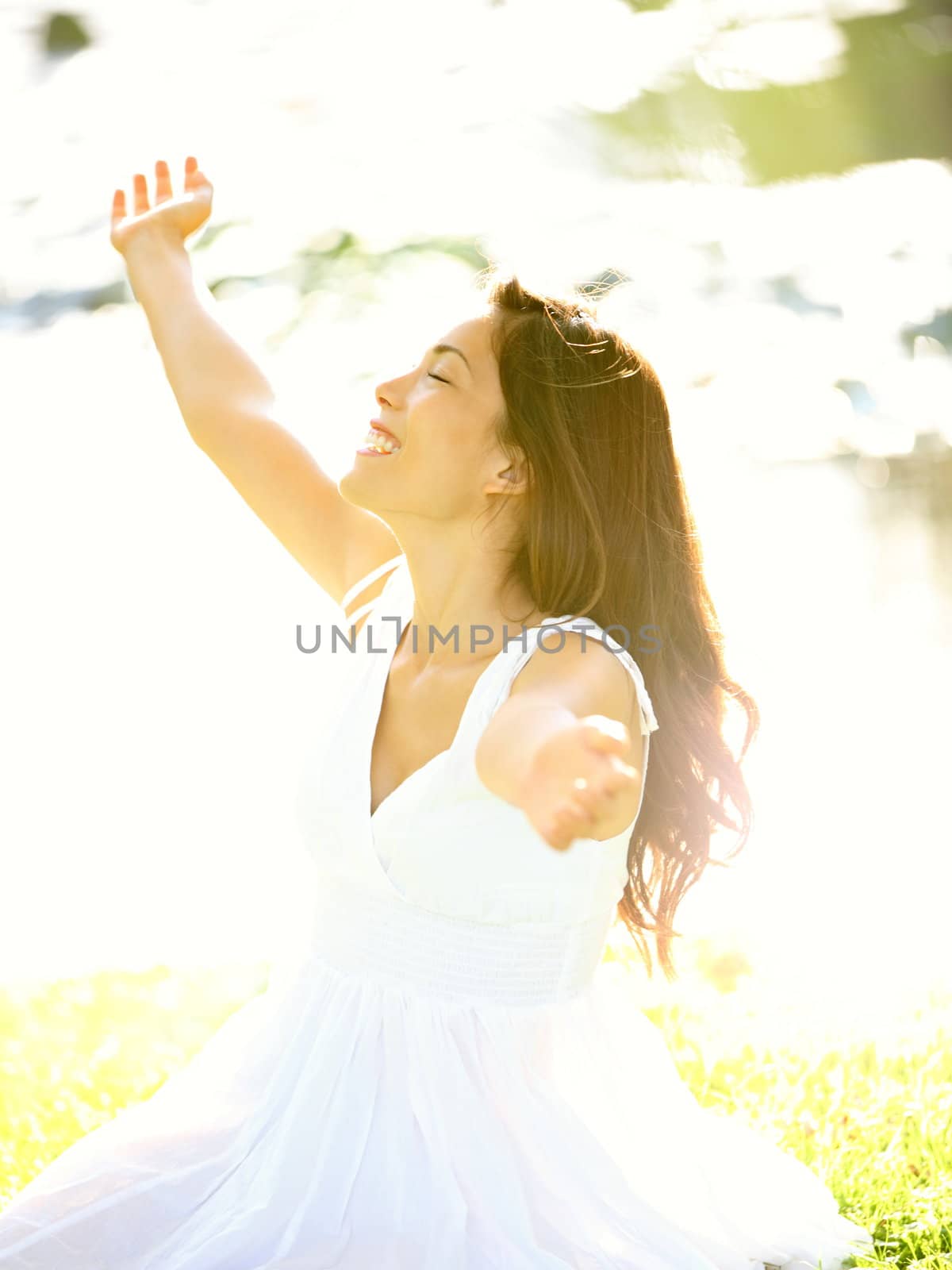 Happy free woman carefree in spring or summer with arms out enjoying the sun joyful in white summer dress sitting in the grass by lake in park. Beautiful mixed race Caucasian / Asian Chinese girl outdoors.