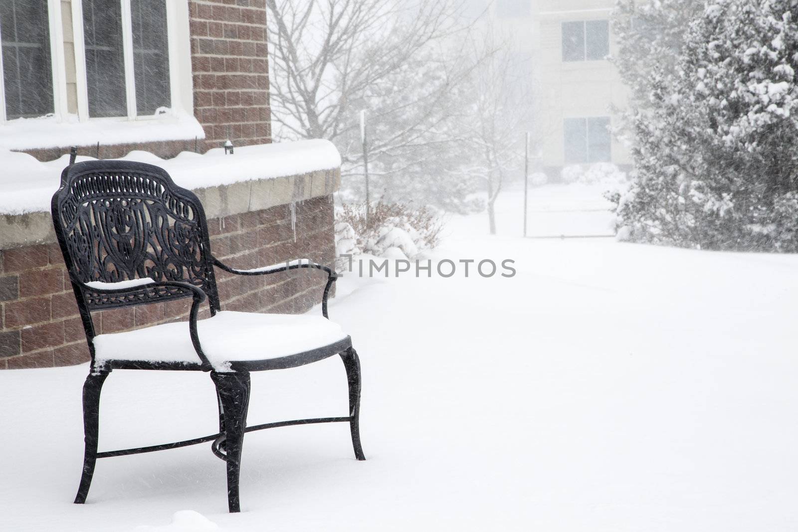 An empty, black, wrought iron bench in a snow storm by a brick building