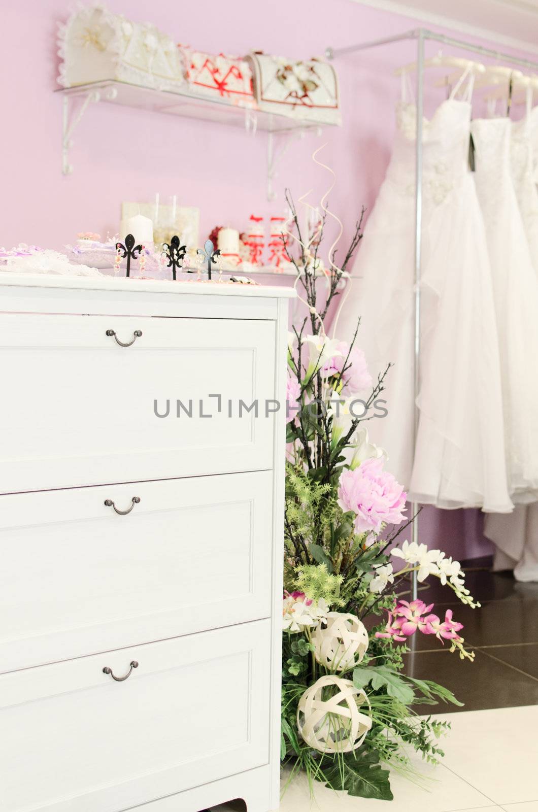 Wedding store background. White bridal dresses. Artificial flowers and different stuff on glass shelfs