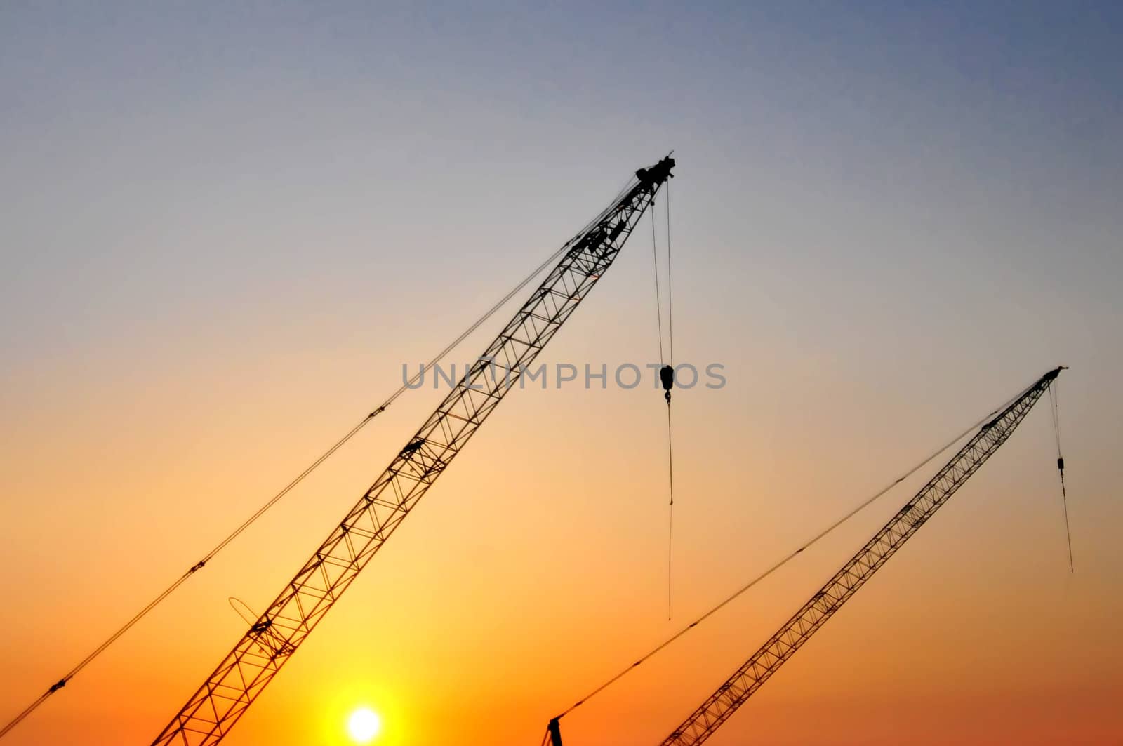 Industrial construction cranes and building silhouettes over sun at sunrise.