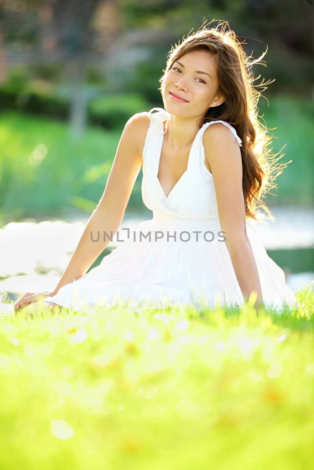 Summer / spring woman smiling happy sitting in white dress in sunny park by lake. Beautiful mixed ethnic Caucasian / Chinese Asian girl outdoor.