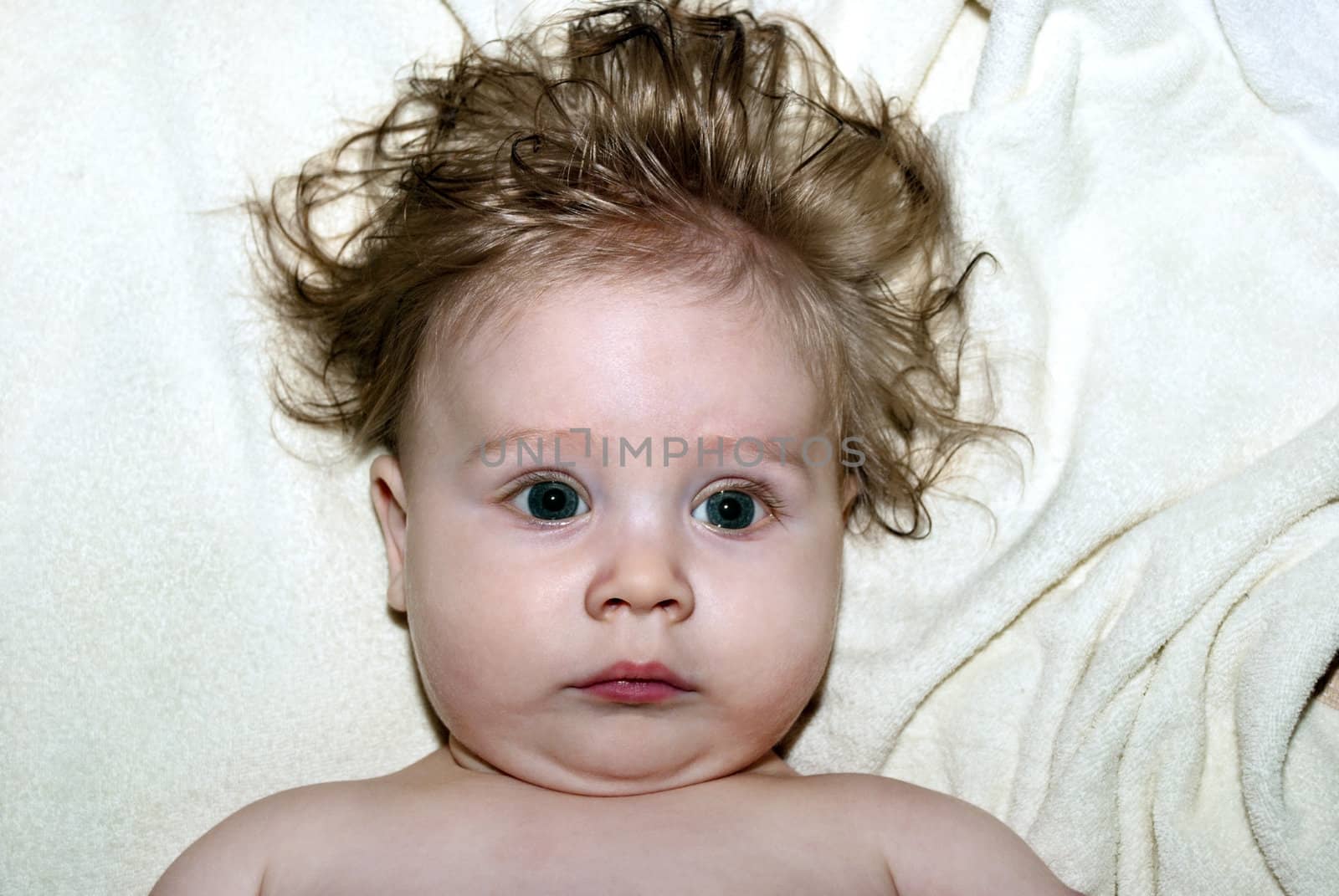 Cute little kid with shaggy hair, lying on the bed looking at the camera.
