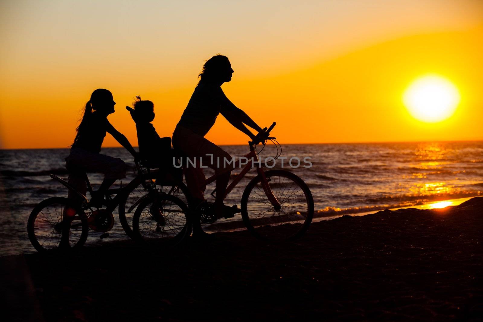 Mother and her kids on the bicycle silhouettes on beach at sunset