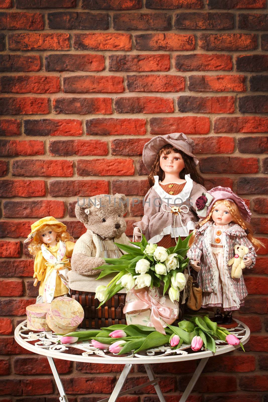 Some porcelain dolls and teddy bears on a table against a wall from a red brick