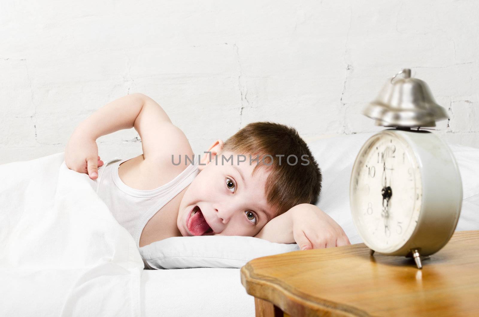 Small toddler boy (4 years old) is lying in bed and showing tongue. He is looking at the camera. Old clock show 6 o'clock. Brick wall