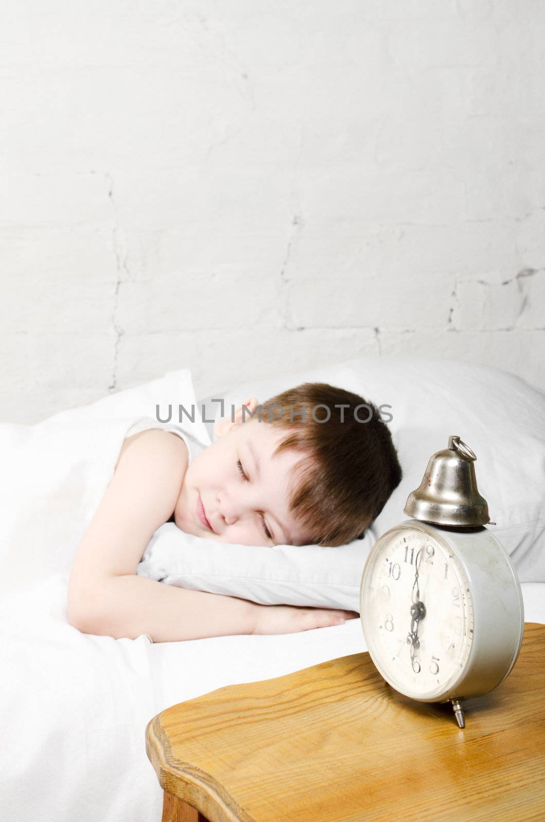 Small toddler boy (4 years) is sleeping in bed. Brick wall at the background. Old clock show 6 o'clock.