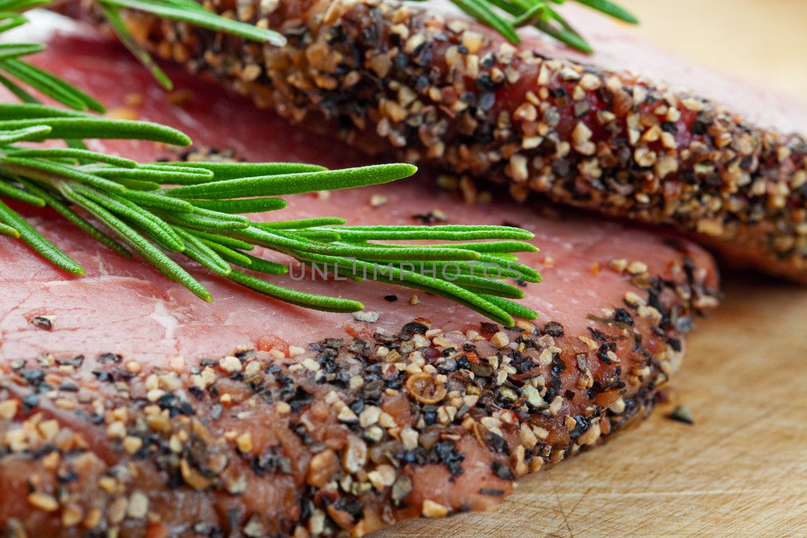 Two raw, eye of round, Alberta beef steaks, encrusted with pepper & spices and topped with fresh rosemary.  BBQ ready!  Shallow depth of field.