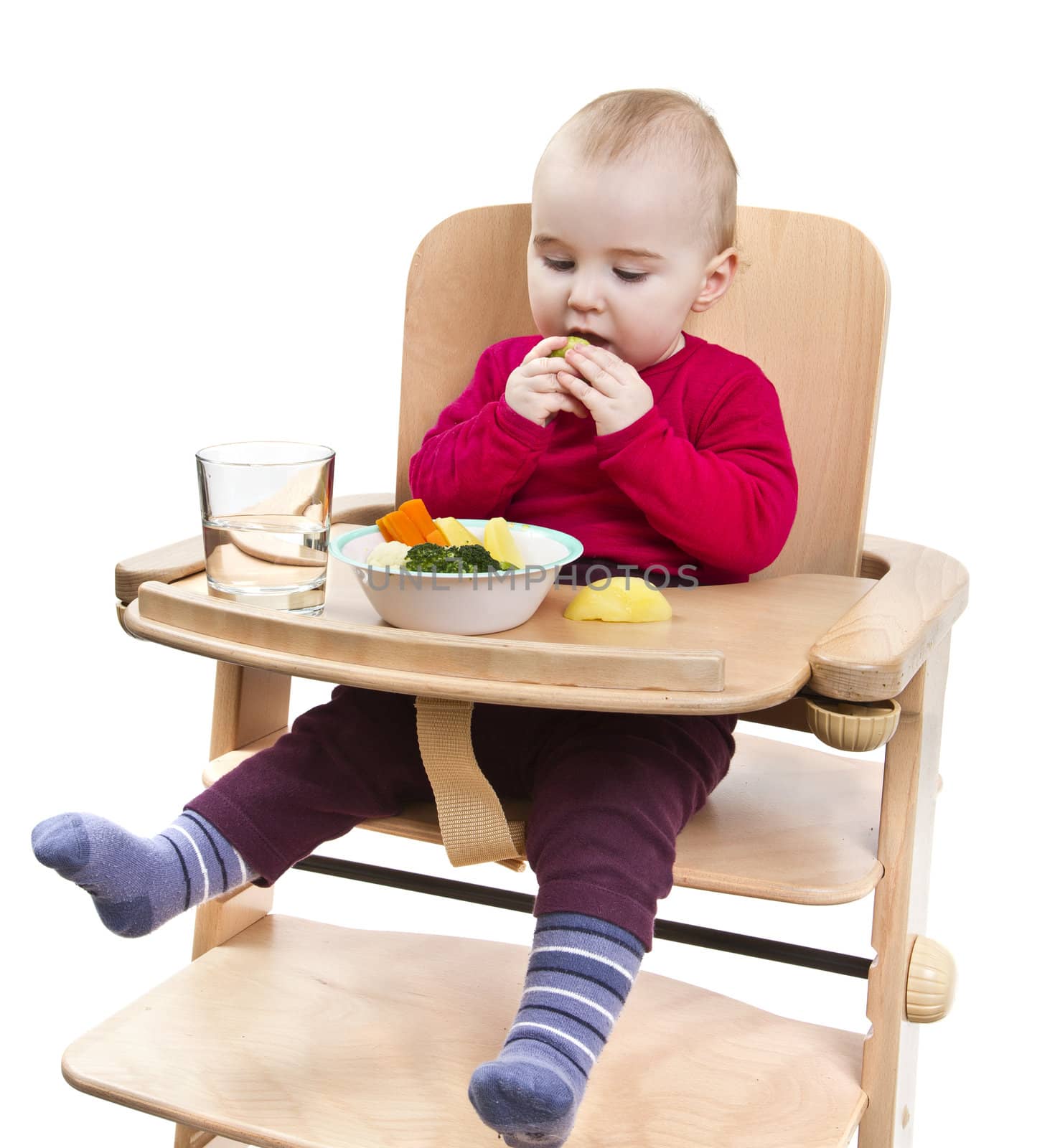 young child in red shirt eating vegetables in wooden chair. Isolated on white background