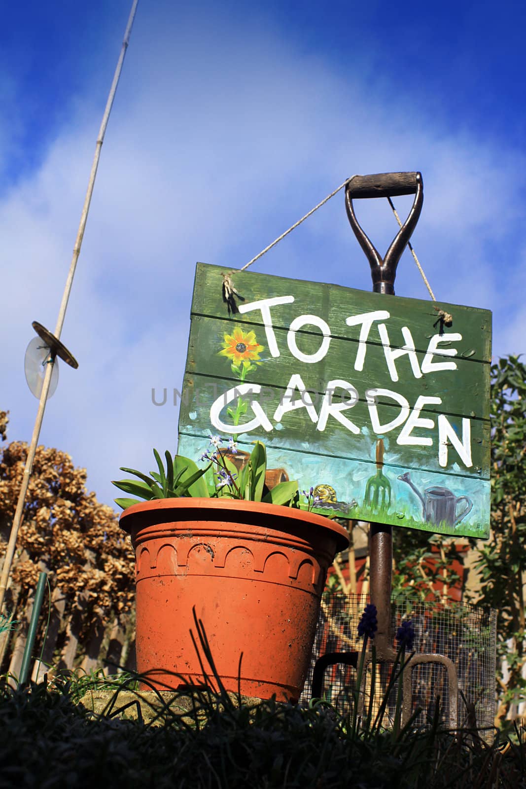 A portrait format image set at a low angle, of a terracotta colored flower pot with a garden fork and a hand painted wooden sign with garden themed images. Set in a small city garden against a bright blue sky background.