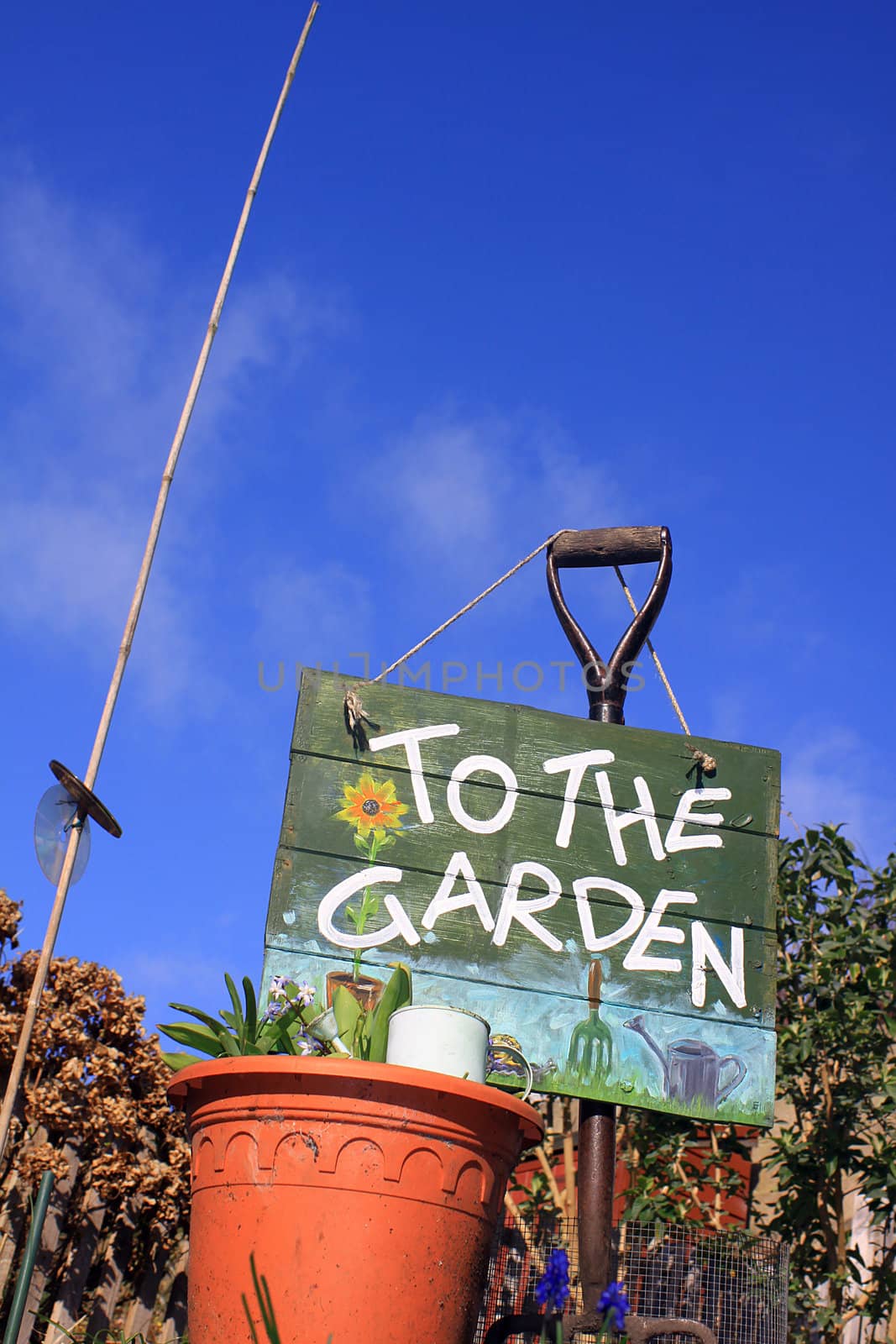 A portrait format image set at a low angle, of a terracotta colored flower pot set in front of a garden fork with a hand painted sign with garden themed images. Set in a small city garden against a bright blue sky background.