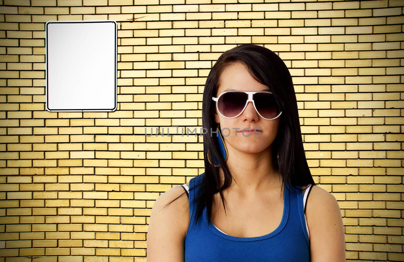 Tough looking young woman with sunglasses in front of yellow brick wall background with blank white metal sign ready for your text.