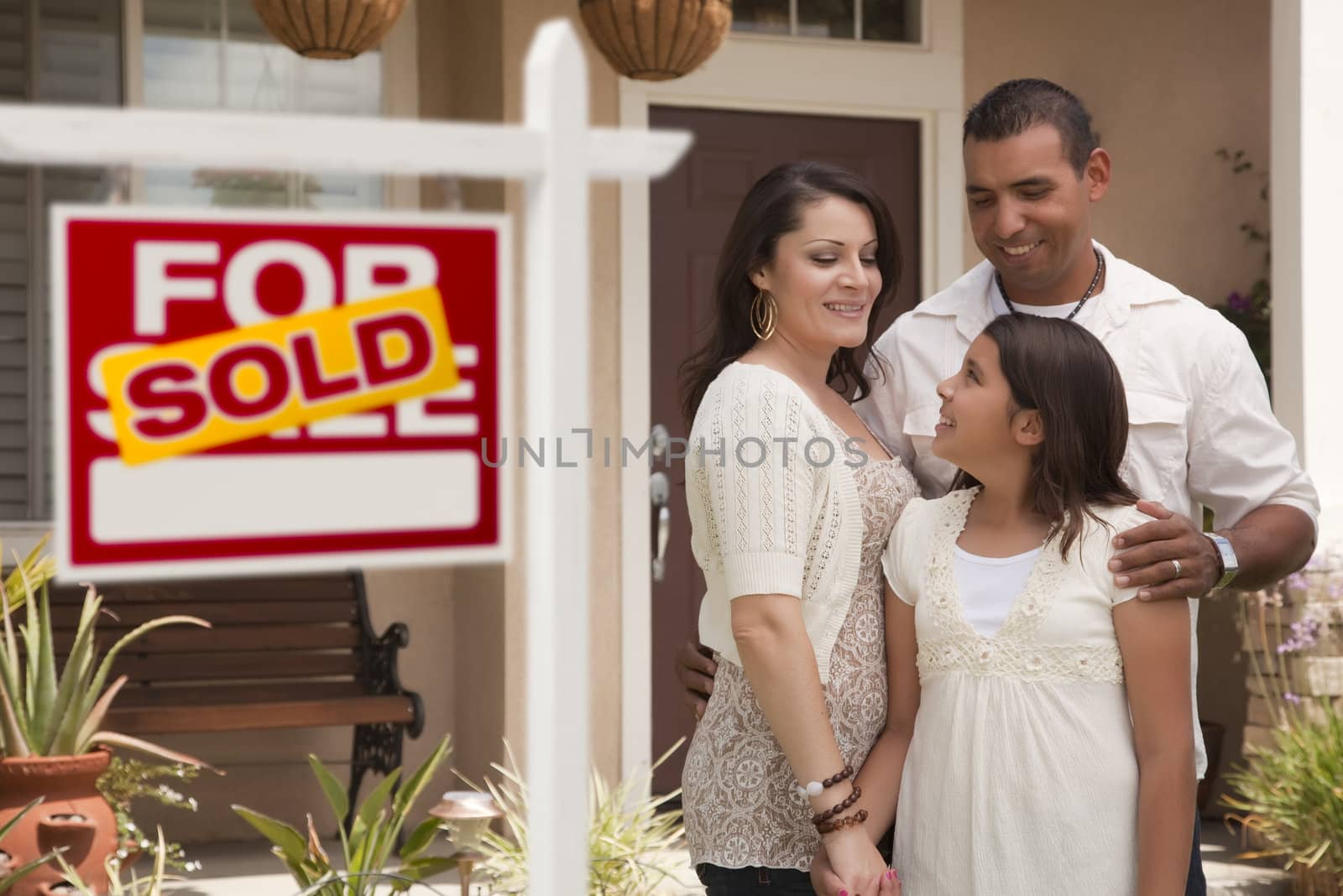 Hispanic Mother, Father and Daughter in Front of Their New Home with Sold Home For Sale Real Estate Sign.