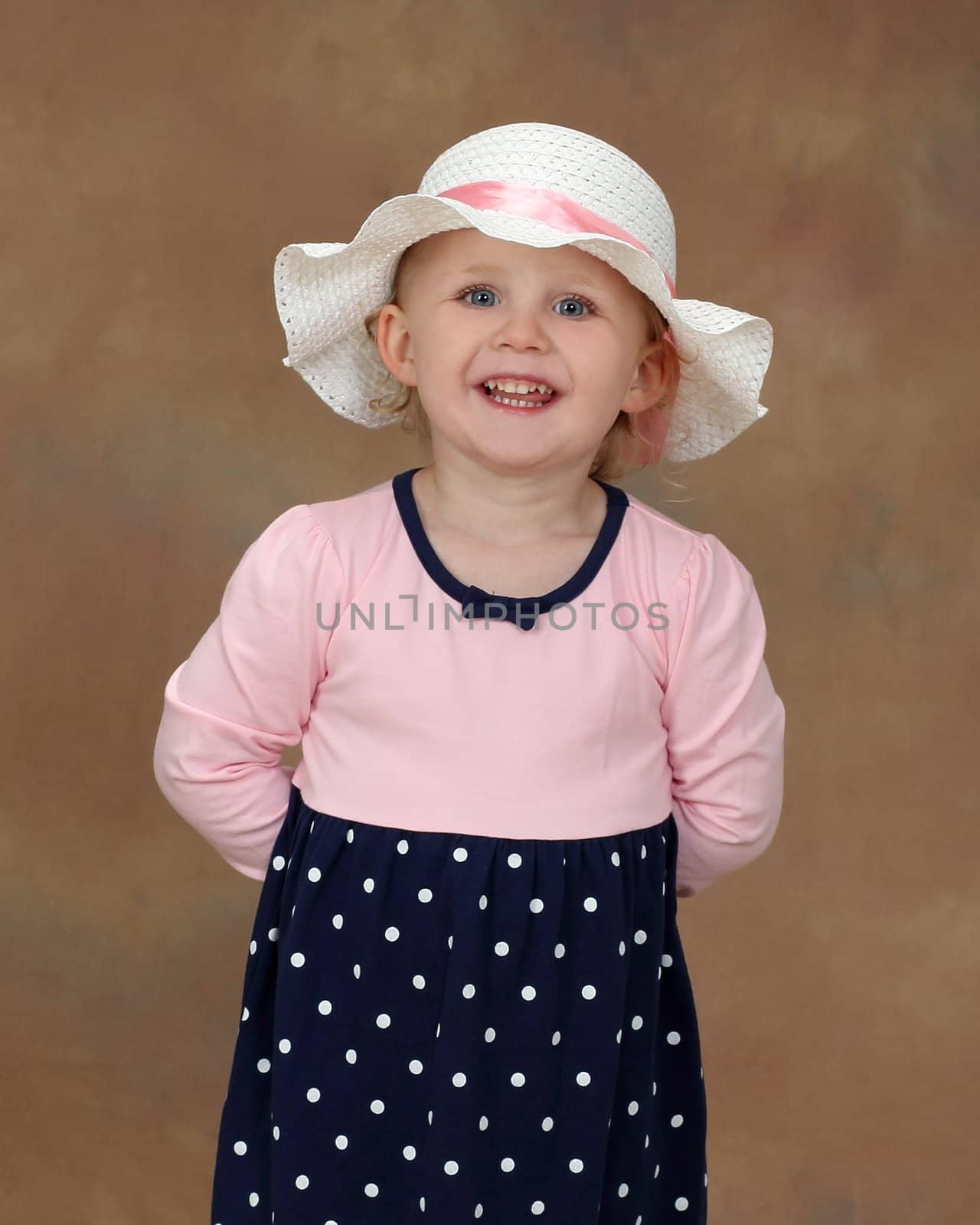 A little blond girl with blue eyes and summer hat smiles happily at the camera