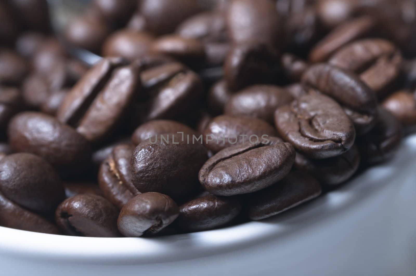 Close-up (macro) of coffee beans piled into a white china coffee cup.  Shallow depth of field with silver spoon just visible in soft focus.