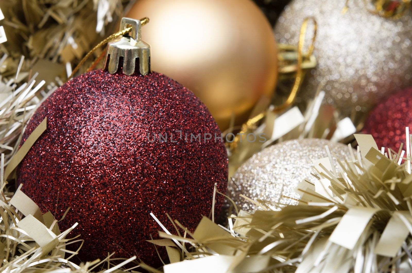 Close up of Christmas baubles resting on and surrounded by a pale gold tinsel garland.  Sparkly red bauble has focus in foreground.  Matte gold and sparkly silver varieties in background.  Shallow depth of field.