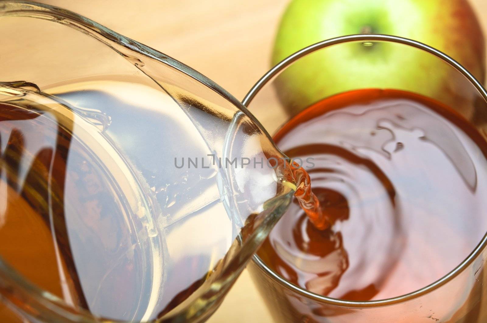 Close up of apple juice being poured from a glassware jug into a glass over wooden table.  Apple in top right frame.