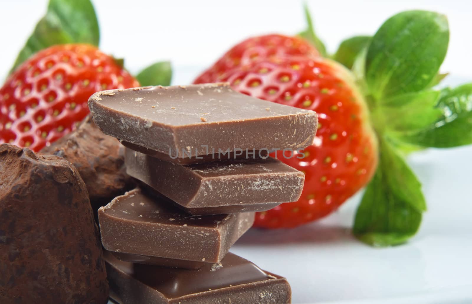 Close up of a stack of chocolate pieces with dusted truffles to the left, and strawberries in soft focus in the background.  The surface is a grey-blue china plate.