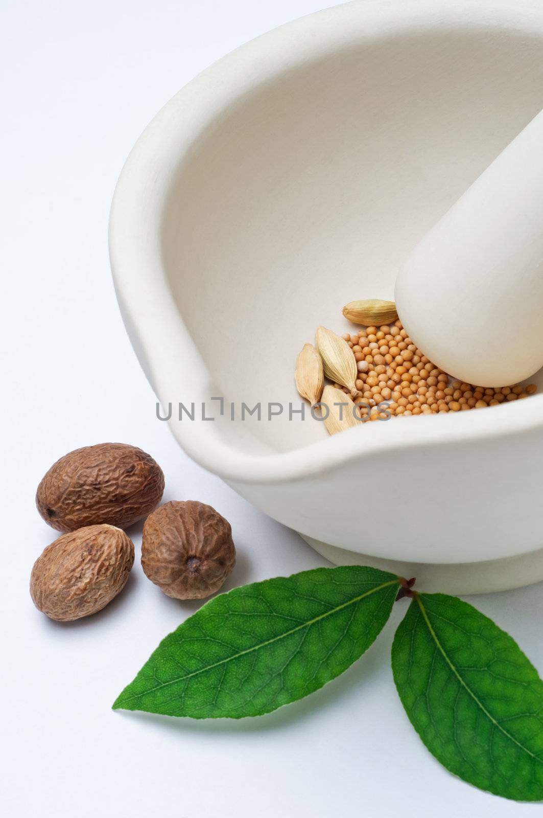 Close up of a stoneware pestle and mortar containing a mixture of whole spices, with nutmegs and bay leaves on the surface outside it. Vertical (portrait) orientation.