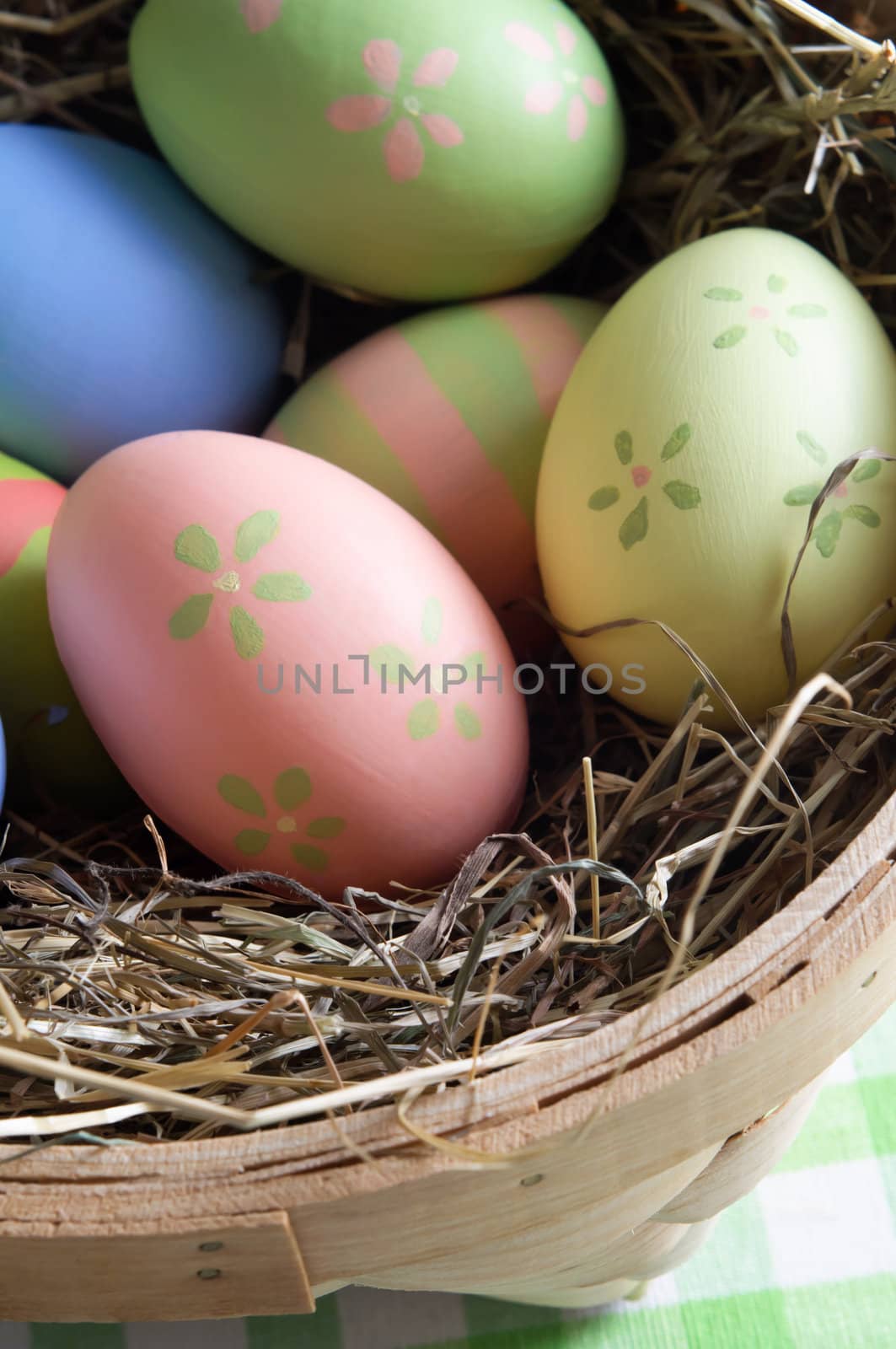 Close up of colourful, hand painted Easter eggs nestling in straw inside a basket on a gingham table cloth. Shot from above.