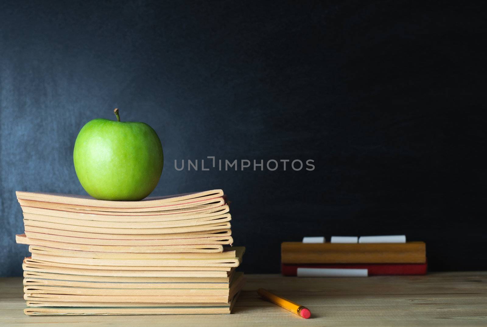 A school teacher's desk with stack of exercise books and apple in left frame. A blank blackboard in soft focus background provides copy space.