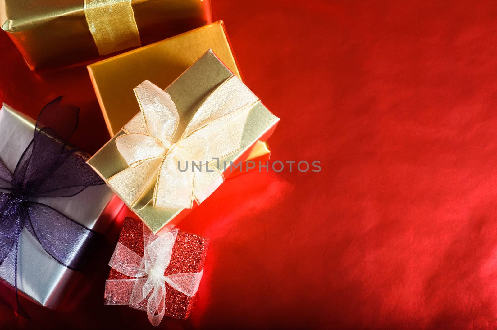 An overhead, horizontal shot of Christmas gift boxes, wrapped and tied with ribbon bows.  Copy space provided to the right on a reflective red background. 