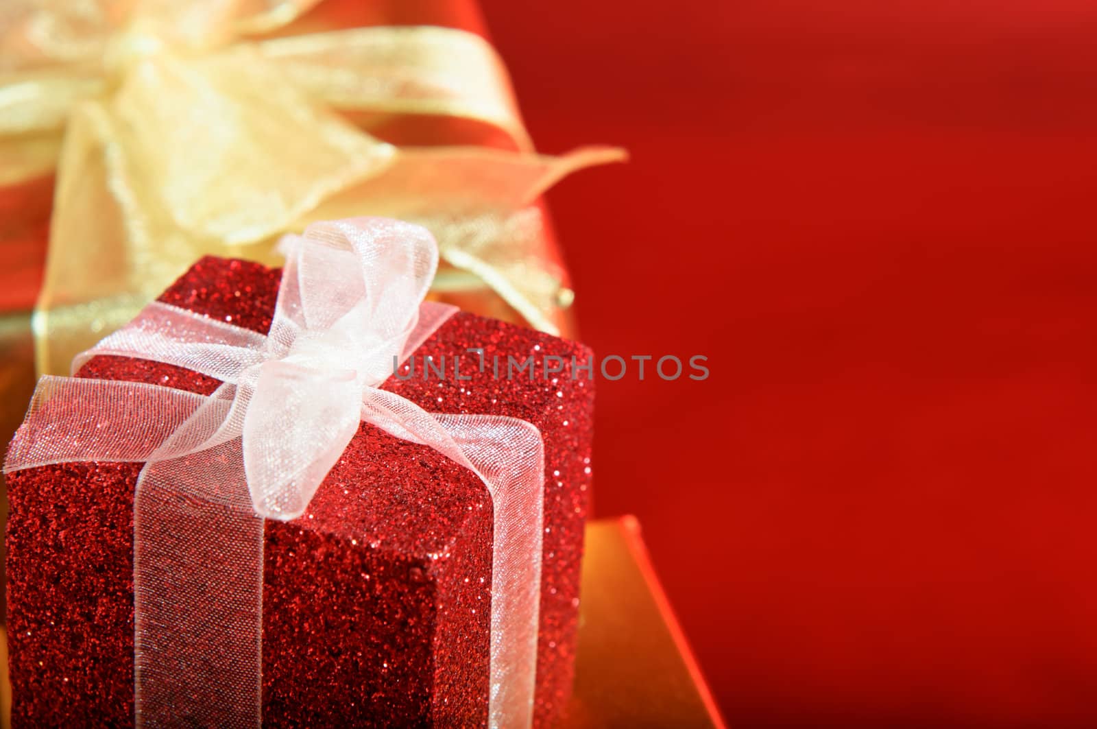 Close up of a glittery red gift box with white ribbon.  Gold box and bow in the background.  Red copy space to right.