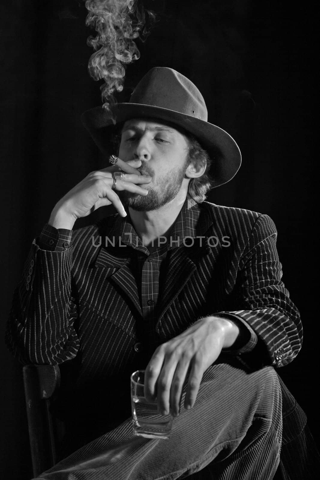 A handsome man in a hat sitting with a glass of alcohol and cigar